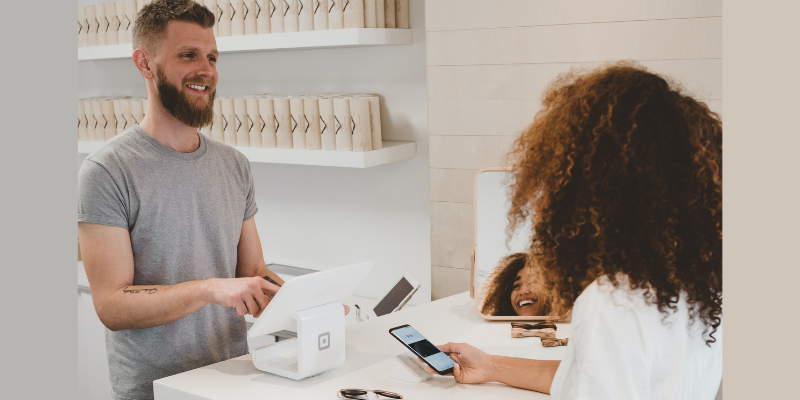Woman paying at a cash register with her phone, while man is running the register at his small business